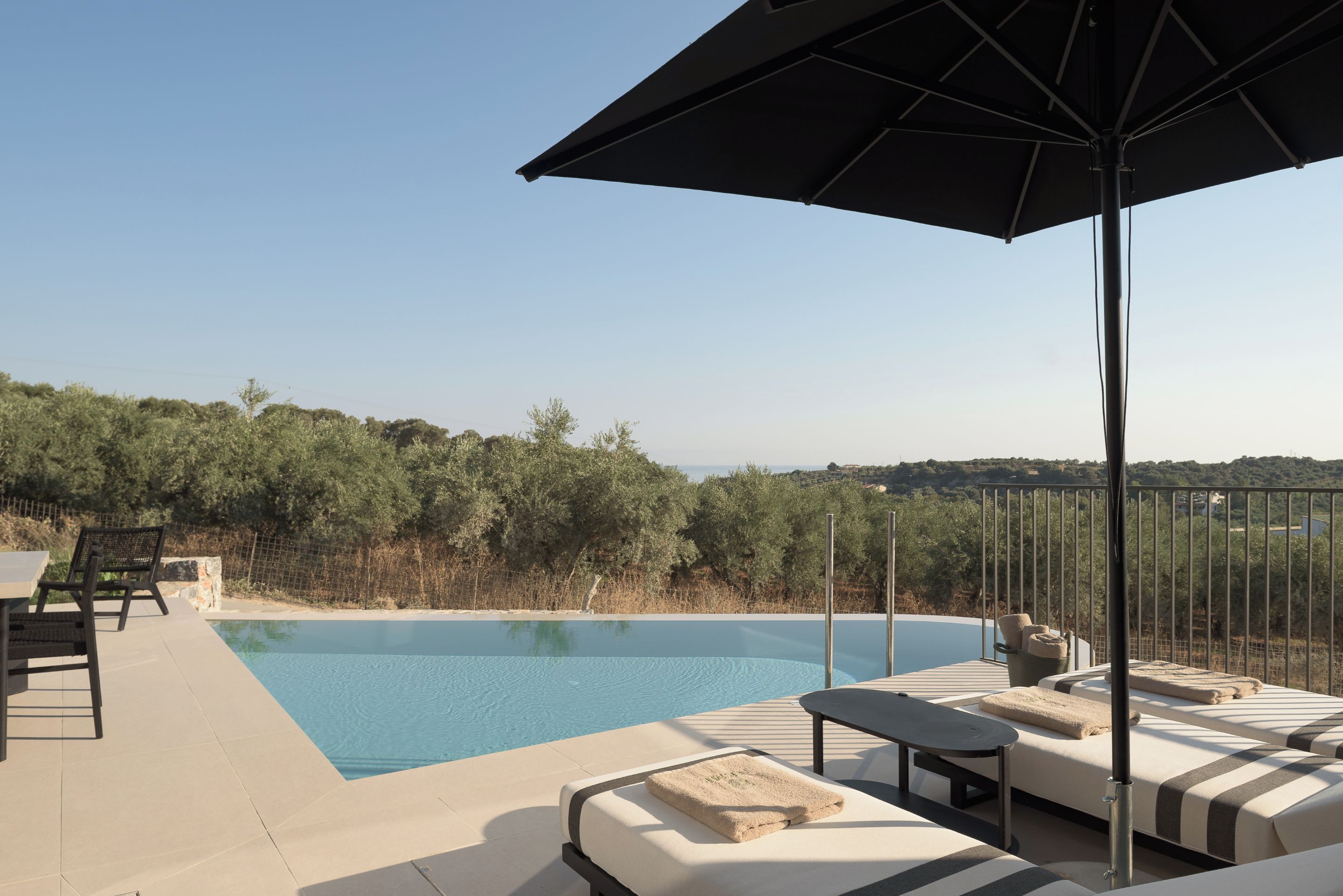 Zona junto a la piscina con sillones de descanso y una sombrilla, con vistas a una masa de agua y una ladera boscosa.