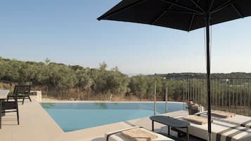 Zona junto a la piscina con sillones de descanso y una sombrilla, con vistas a una masa de agua y una ladera boscosa.