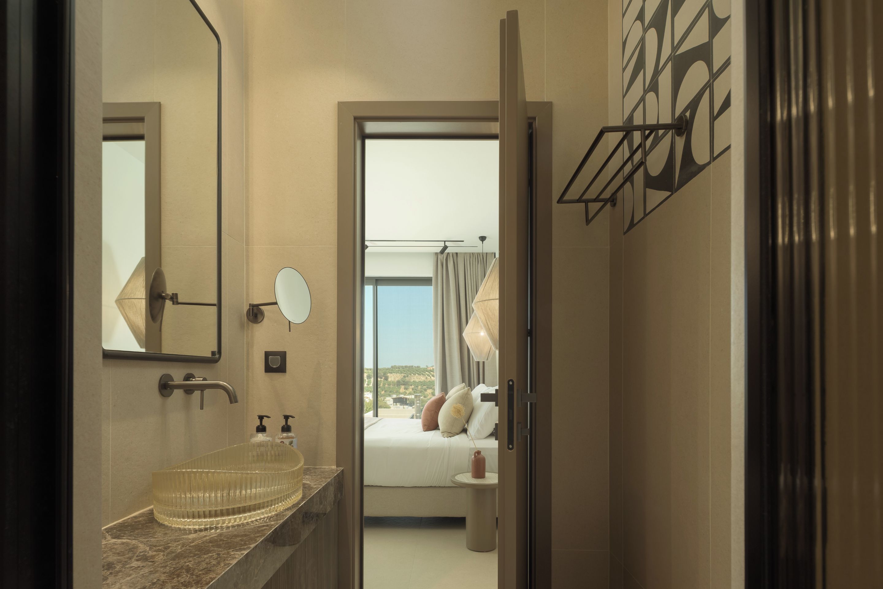 Une salle de bain moderne avec un lavabo en verre, un grand miroir et une vue sur une chambre à coucher avec un lit et une fenêtre.
