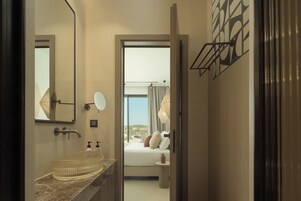 Un baño moderno con un lavabo de vidrio, un espejo grande y vistas a un dormitorio con cama y ventana.