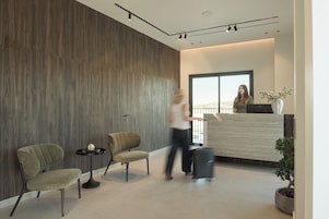 A modern reception area with a wooden wall, two upholstered chairs, a small table with a book, and a potted plant.