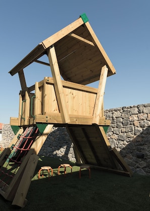 Une structure de jeu en bois, dotée d’un mur d’escalade et de toboggans, installée devant un mur de pierre.