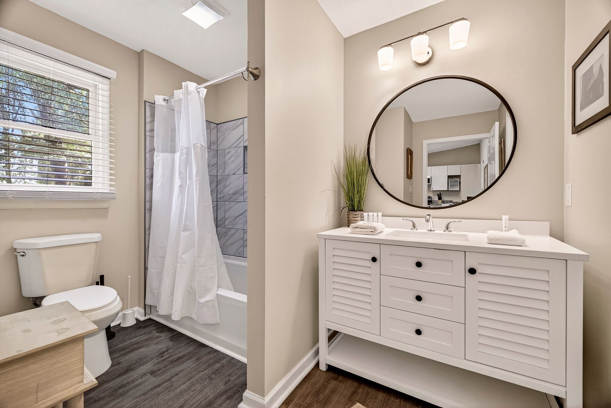 A bathroom with a toilet, shower, vanity with a mirror, and a window with blinds.