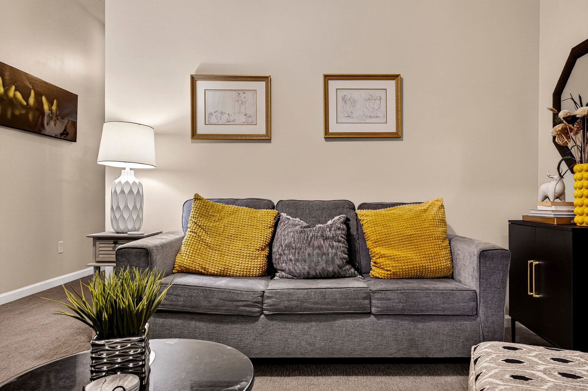 A living room with a grey sofa, yellow and grey cushions, a lamp, framed artwork, and a plant on a table.
