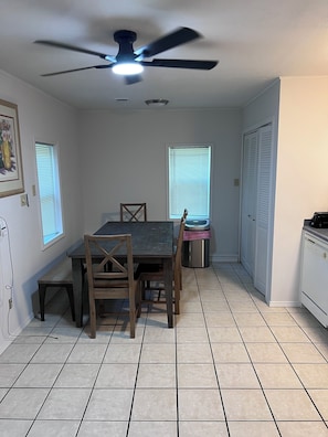 A dining area with a table and chairs, a ceiling fan, a trash can, and a kitchen area with cabinets.