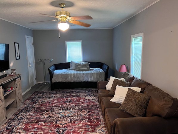 A bedroom with a bed, a brown leather sofa, a patterned rug, a ceiling fan, and two windows with blinds.