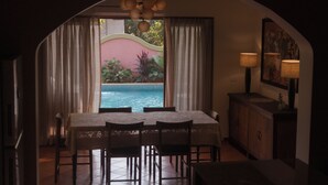 A dining area with a view of a pool, a table with chairs, and a cabinet with a lamp. - Flor De Lis - 4 BHK Villa w/Pool, wifi & parking behind Sakana in Anjuna (Vagator)