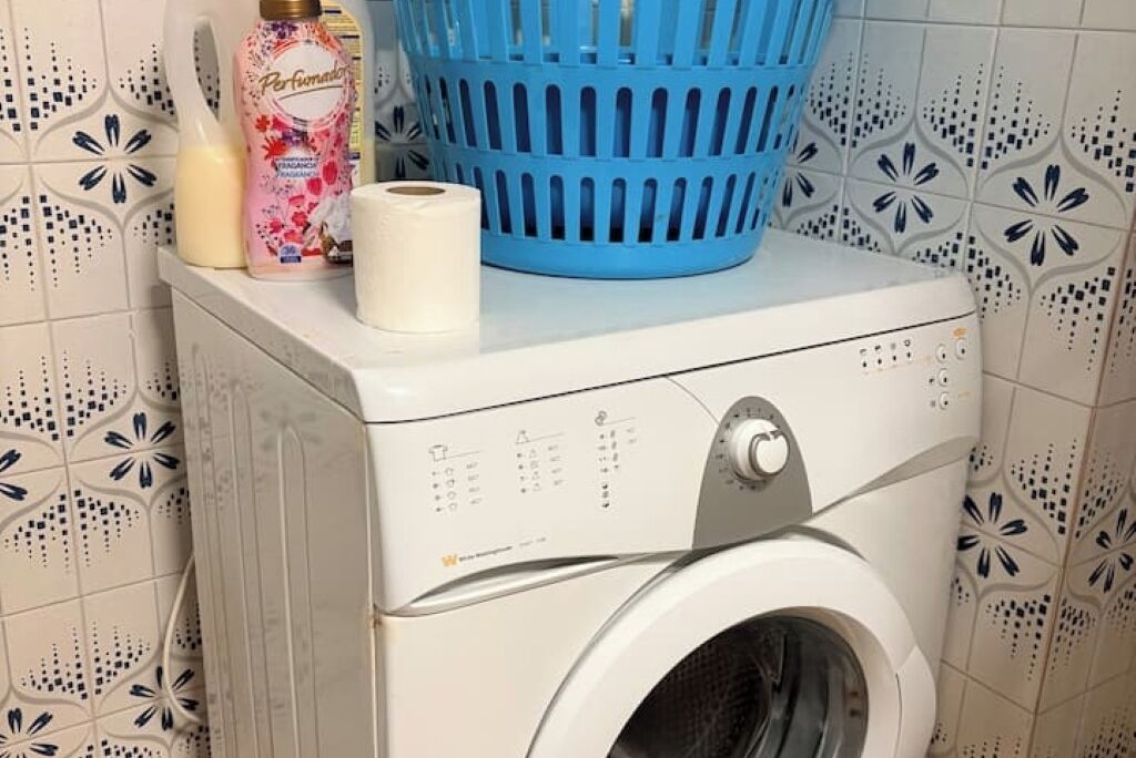 Eine Waschmaschine mit Frontlader, ein blauer Wäschekorb darauf, eine Waschmittelflasche und eine Toilettenpapierrolle.