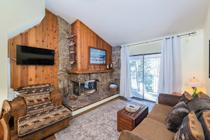 A cozy living room with a stone fireplace, wooden walls, and a large window offering a view of trees. - Newly renovated 2 bedroom, vaulted ceilings, hardwood floor and more! (Winter Park)