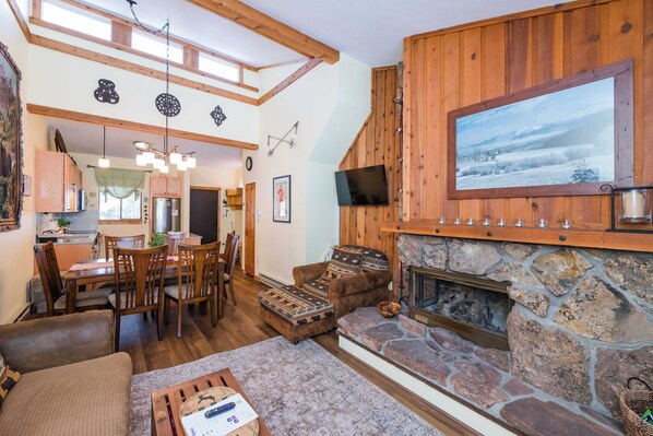 A cozy living room with a stone fireplace, wooden walls, and a dining area with a high ceiling and exposed beams. - Beaver Village Unit #0233 Renovated 2 bed vaulted ceilings, hardwood floor (Winter Park)