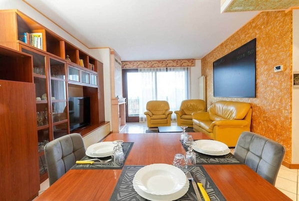A dining area with a wooden table set for two, flanked by leather chairs. A bookshelf with glass doors is on the left, and a yellow sofa is on the right.