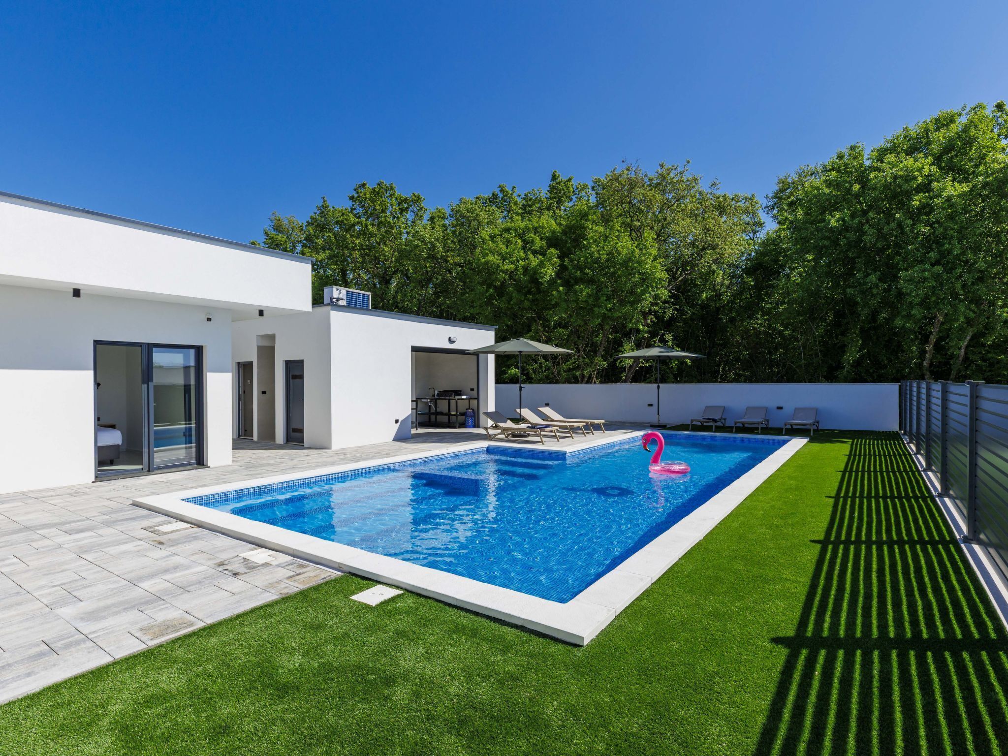 A modern pool area with a clear blue pool, a pink flamingo float, and a white building with a glass door entrance.
