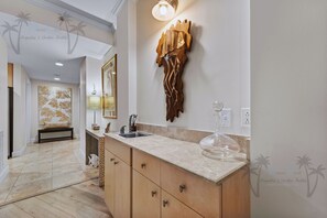A modern bathroom with a marble countertop, wooden cabinets, a large mirror, and a decorative wall piece. - Sea Breeze 601 | Condo with Beach Access! (Biloxi)