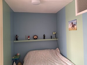 Un dormitorio con una cama, un estante con figuritas, un escritorio con lámpara y un cuadro en la pared.