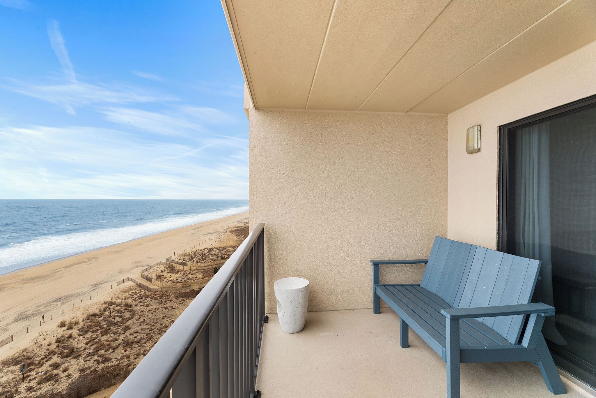 A balcony with a view of the beach, a white trash can, and a blue bench.