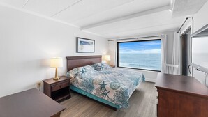 Ein Hotelzimmer mit Bett, Nachttischen, einem Schreibtisch, einem Fenster mit Meerblick und einem gerahmten Bild an der Wand.