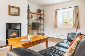A cozy living room with a fireplace, a wooden table, a black leather sofa, and a window with curtains. - 2 Ruscon Cottage,  Brancaster Staithe, Norfolk (Brancaster Staithe)