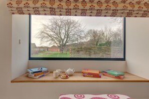A window seat with a view of trees and a house, a wooden shelf with books and a teapot, and a patterned curtain above. - Pheasant Cottage, Little Massingham, Norfolk (Little Massingham)