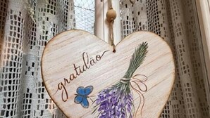 En hjerteformet trædekoration med ordet "gratidão" og et lilla blomstermønster, hængende foran en blondegardin.