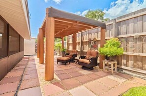 Un patio couvert comprenant deux fauteuils rembourrés et une petite table, le tout entouré d’une clôture en bois et de plantes en pot.