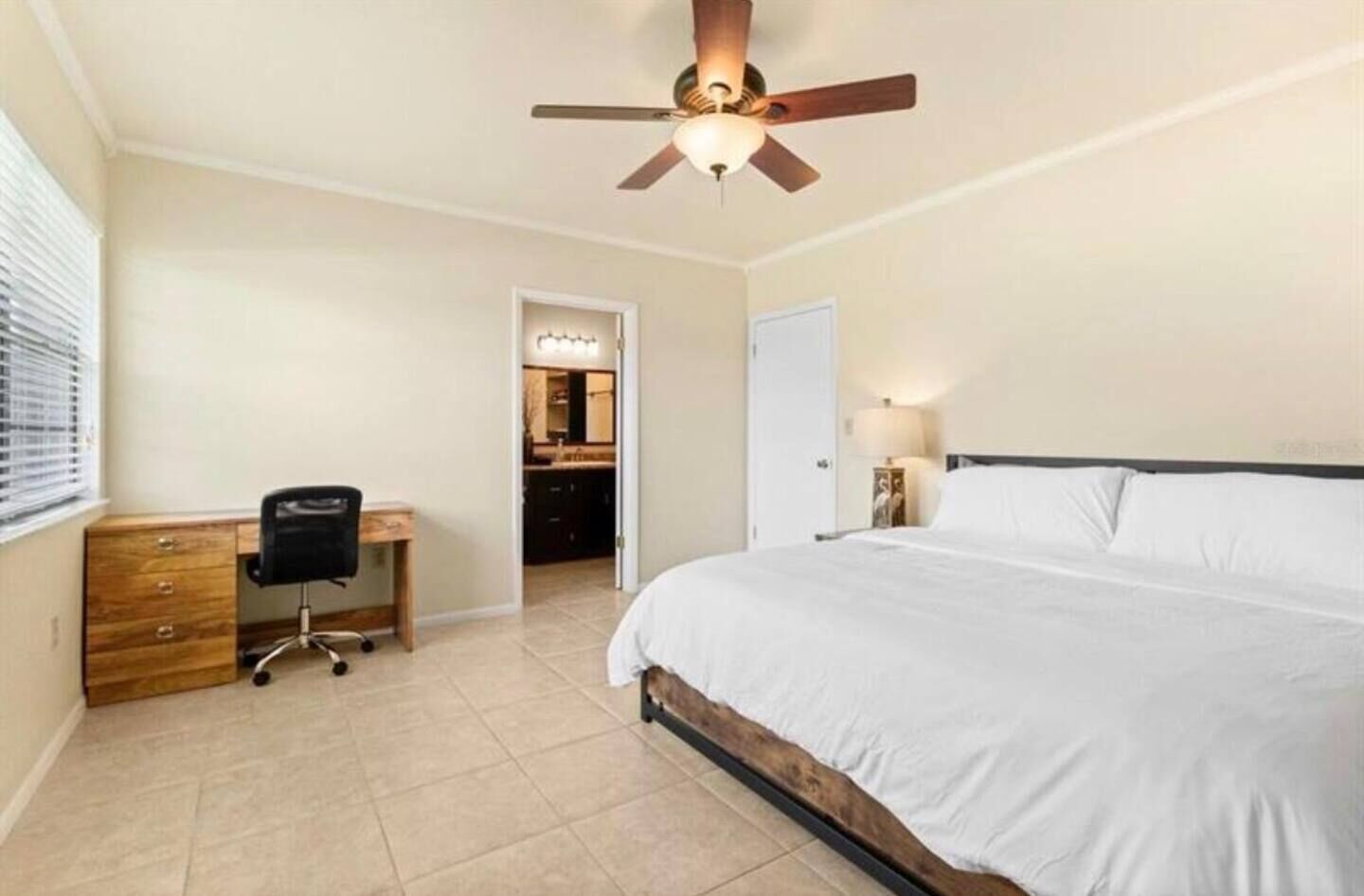 A bedroom with a bed, a desk, a chair, a ceiling fan, and a bathroom visible through an open door.