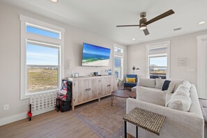 A spacious living room with a large TV, a ceiling fan, and a view of the beach through the windows. - Steele Away by Liquid Life (Gulf Shores)