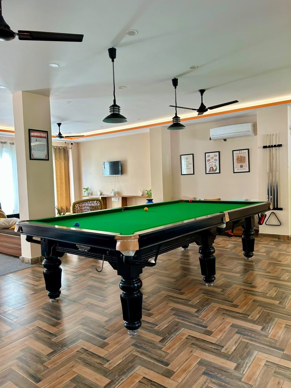 A pool table in a room with wooden flooring, ceiling fans, and framed pictures on the walls.
