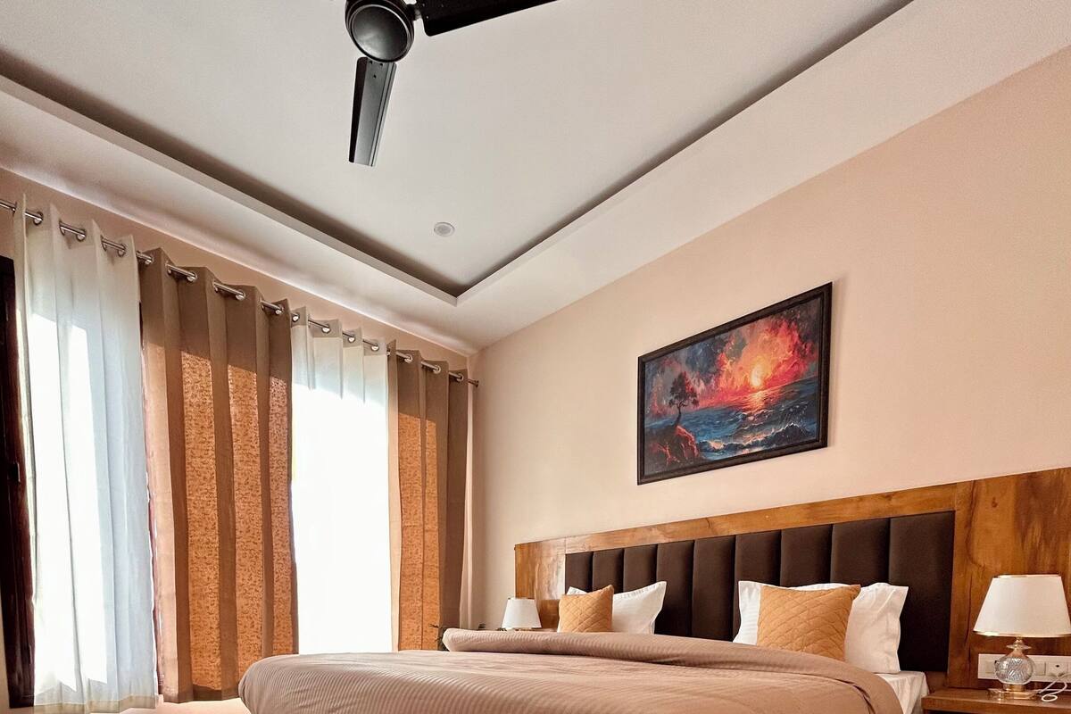 Un dormitorio con una cama grande, un ventilador de techo, un cuadro de un atardecer y una mesita de noche con una lámpara.