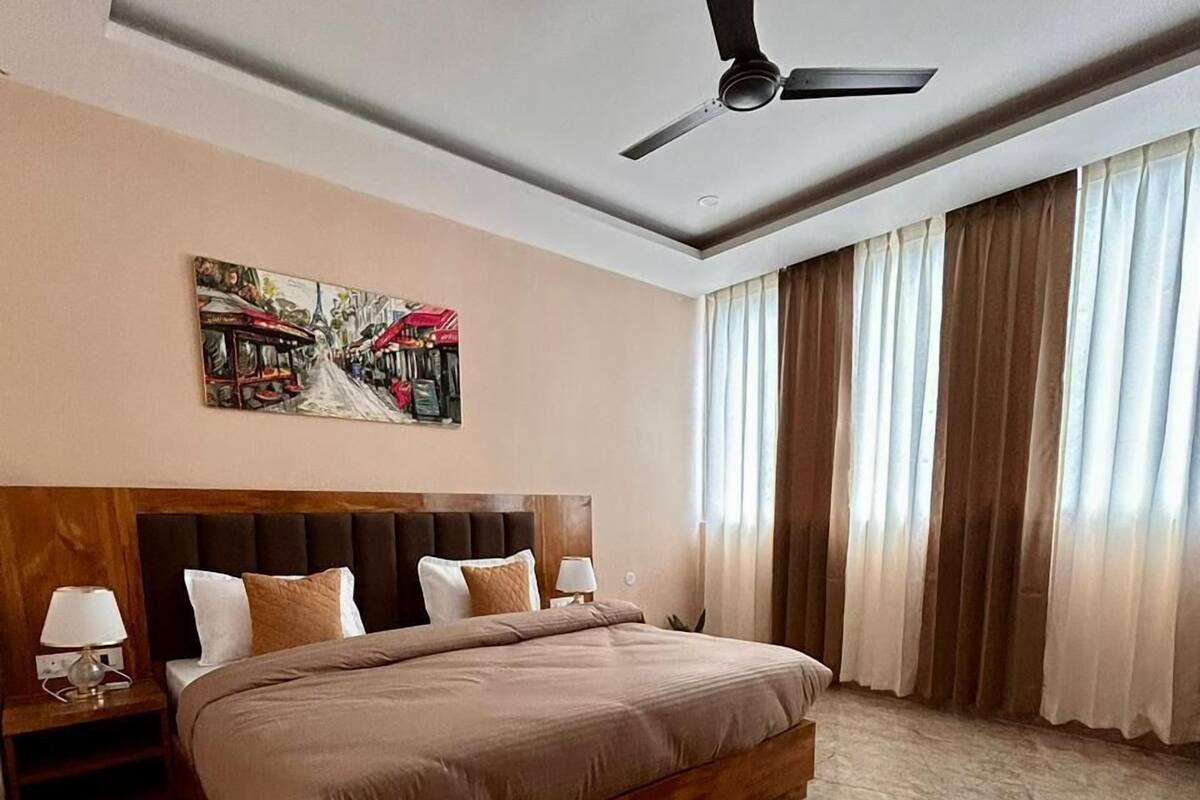 Un dormitorio moderno con una cama grande, un ventilador de techo, un cuadro en la pared y vistas al exterior a través de amplias ventanas.