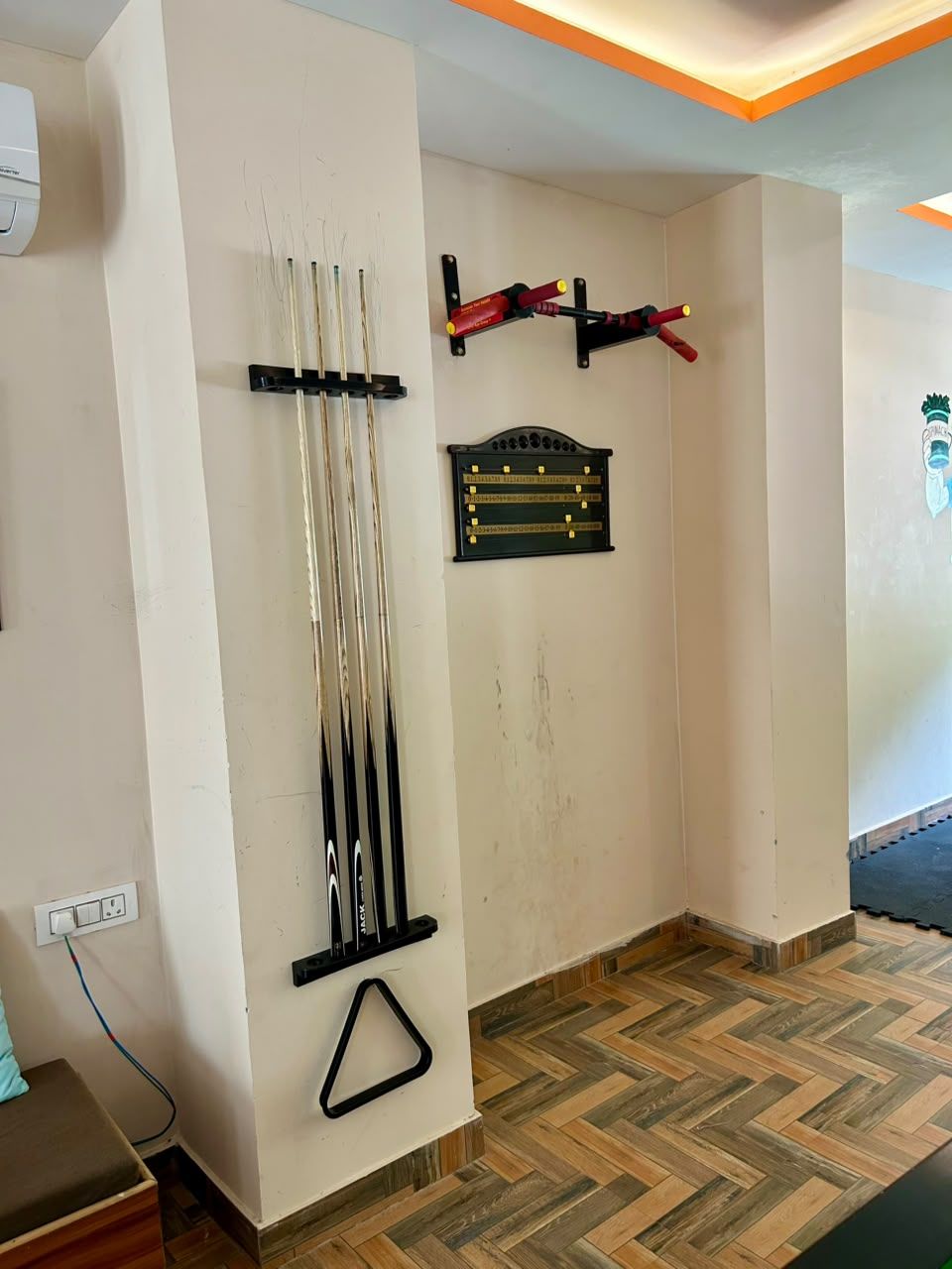 A room with a herringbone-patterned wooden floor, a black triangular decoration on the wall, and a black metal shelf with a plaque.