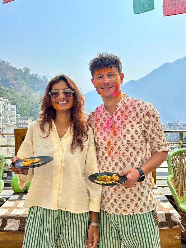 Two people holding plates of food on a terrace with a mountainous backdrop. - Namaste Hostel Rishikesh (Rishikesh)