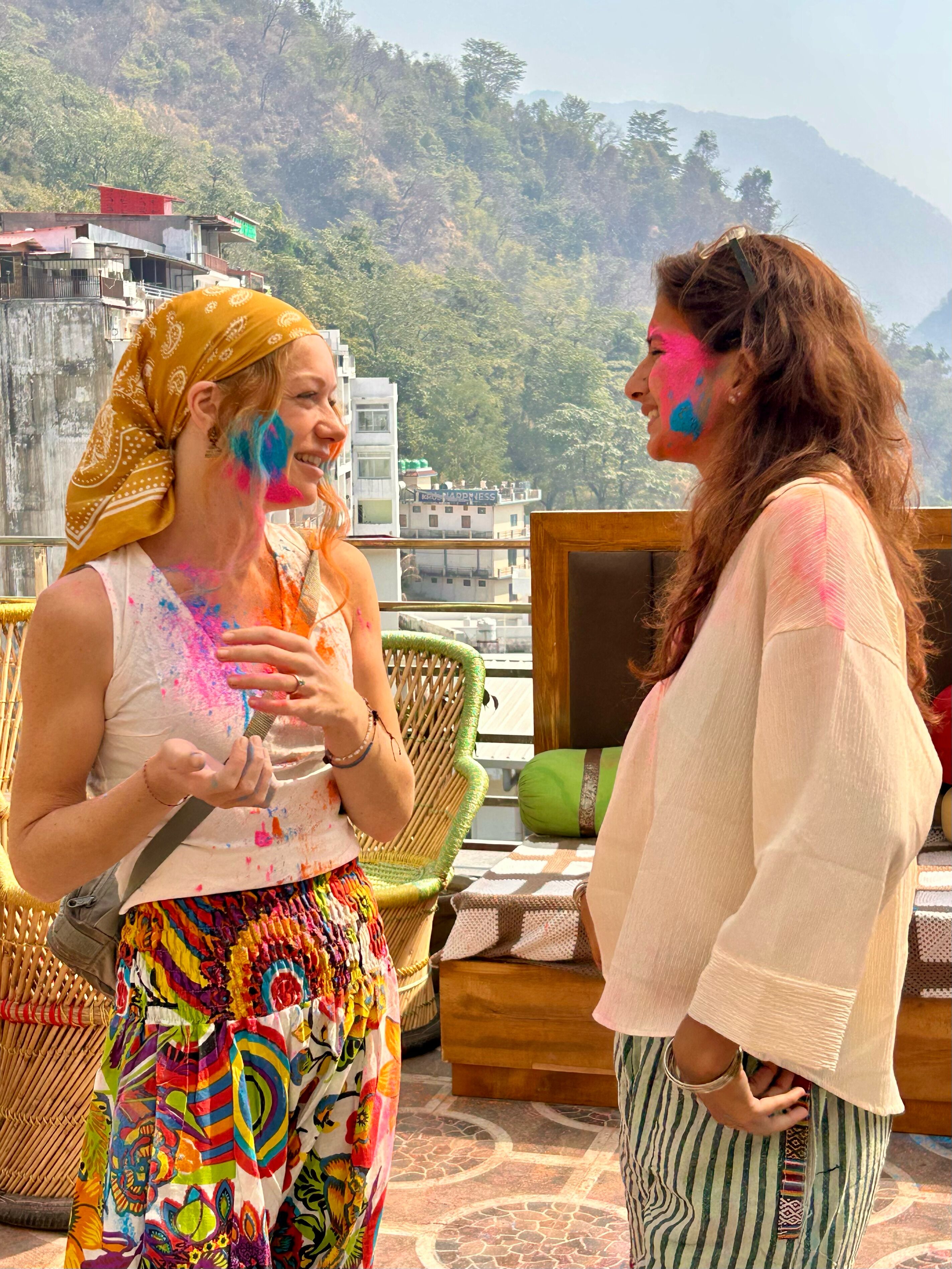 Two people with colorful powder on their faces, standing on a balcony with a view of buildings and hills.