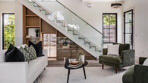 A modern living room with a staircase, a sofa with decorative pillows, a coffee table with books and a plant, and large windows offering views of greenery. - Furnished Modern Luxury Retreat (Austin)