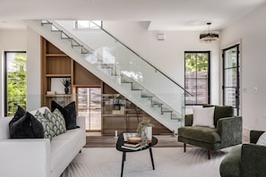 Ein modernes Wohnzimmer mit Treppe, einer Couch mit Zierkissen, einem Couchtisch mit Büchern und einer Pflanze und großen Fenstern mit Blick ins Grüne.