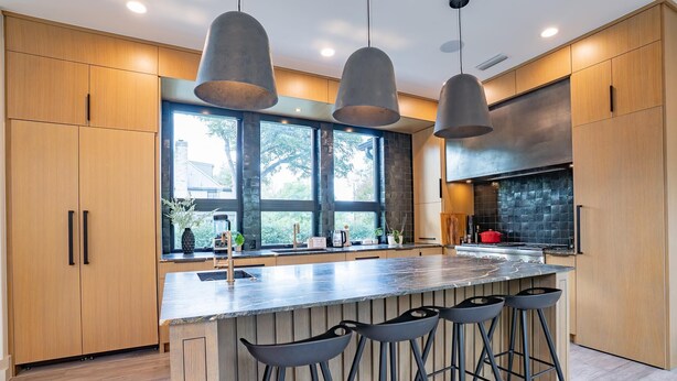 A modern kitchen with wooden cabinets, a large island with bar stools, pendant lighting, and a view through large windows. - Furnished Modern Luxury Retreat (Austin)