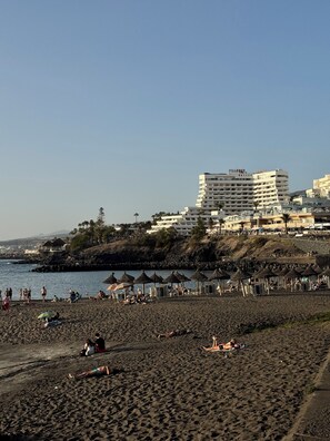 A beach with people sunbathing and swimming, a rocky shoreline, and a large hotel building in the background. - Club Villamar Exclusive Beachfront Penthouse (Costa Adeje)