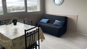 Ein Zimmer mit Esstisch, Stühlen, einer Couch mit blauen Kissen, einer Topfpflanze und einem runden Spiegel an der Wand.