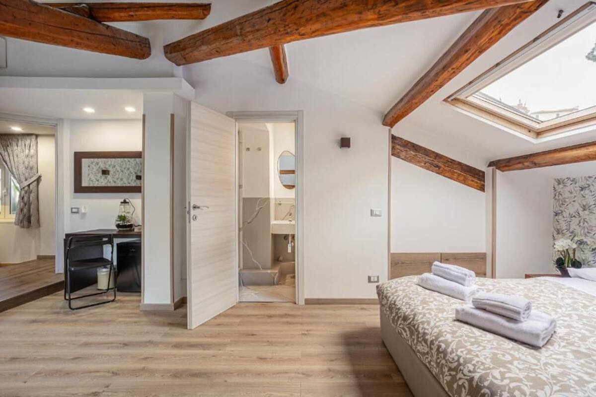 A bedroom with a sloped ceiling, wooden beams, a skylight, a bed with towels, a desk, and a bathroom visible through an open door.