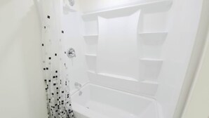 A white bathtub with a shower curtain, a wall-mounted faucet, and a shelf above the tub. - Niagara Cozy Suite - Modern 2bed 15 min to Falls (Thorold)