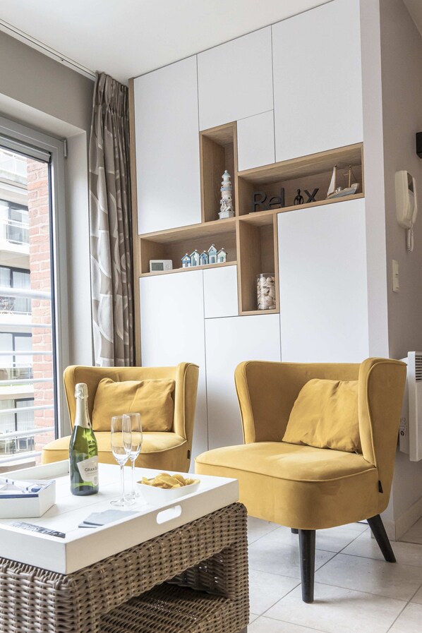 A modern living room with two mustard-colored armchairs, a white coffee table, and a wicker side table. There is a bottle of wine and two glasses on the table. The room has a large window with curtains, a built-in shelving unit with decorative items, and a white wall with a telephone.