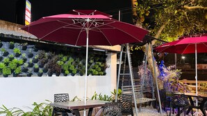 An outdoor seating area with tables and chairs, a red umbrella, and a wall of potted plants. - NAKSHATRA (Neyyattinkara)
