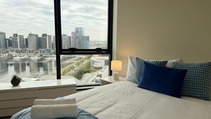 Une chambre d’hôtel dotée d’une grande fenêtre donnant sur la ville, d’un lit soigneusement fait, d’une table de chevet avec une lampe et d’une petite plante posée sur le rebord de la fenêtre.