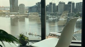 Une vue depuis une fenêtre d’un immeuble de grande hauteur, donnant sur une marina avec des bateaux, un bâtiment moderne remarquable et le profil de la ville.