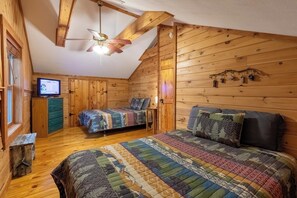 Una habitación acogedora tipo cabaña con paredes de madera, techo inclinado, cama con colcha colorida, escritorio pequeño con computadora y ventana con persianas.