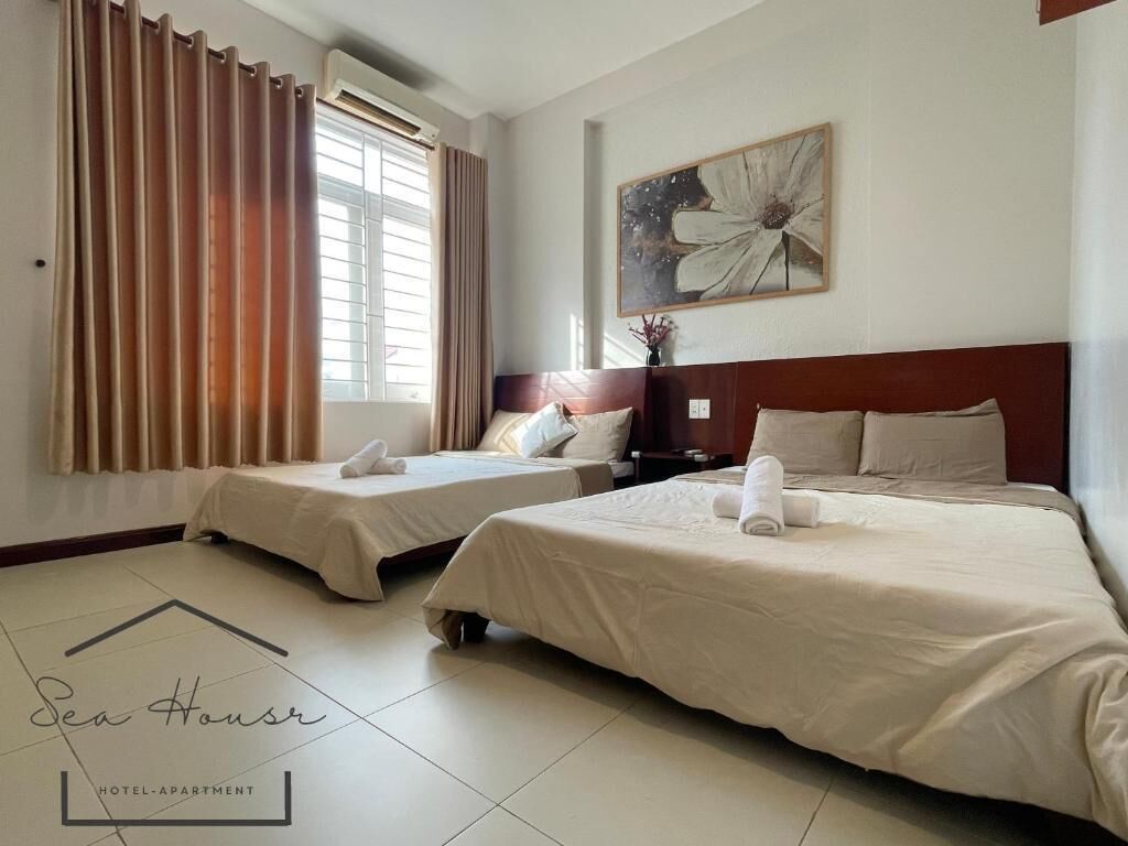 A hotel apartment with two beds, a large window with curtains, a wall-mounted flower painting, and a logo on the floor.