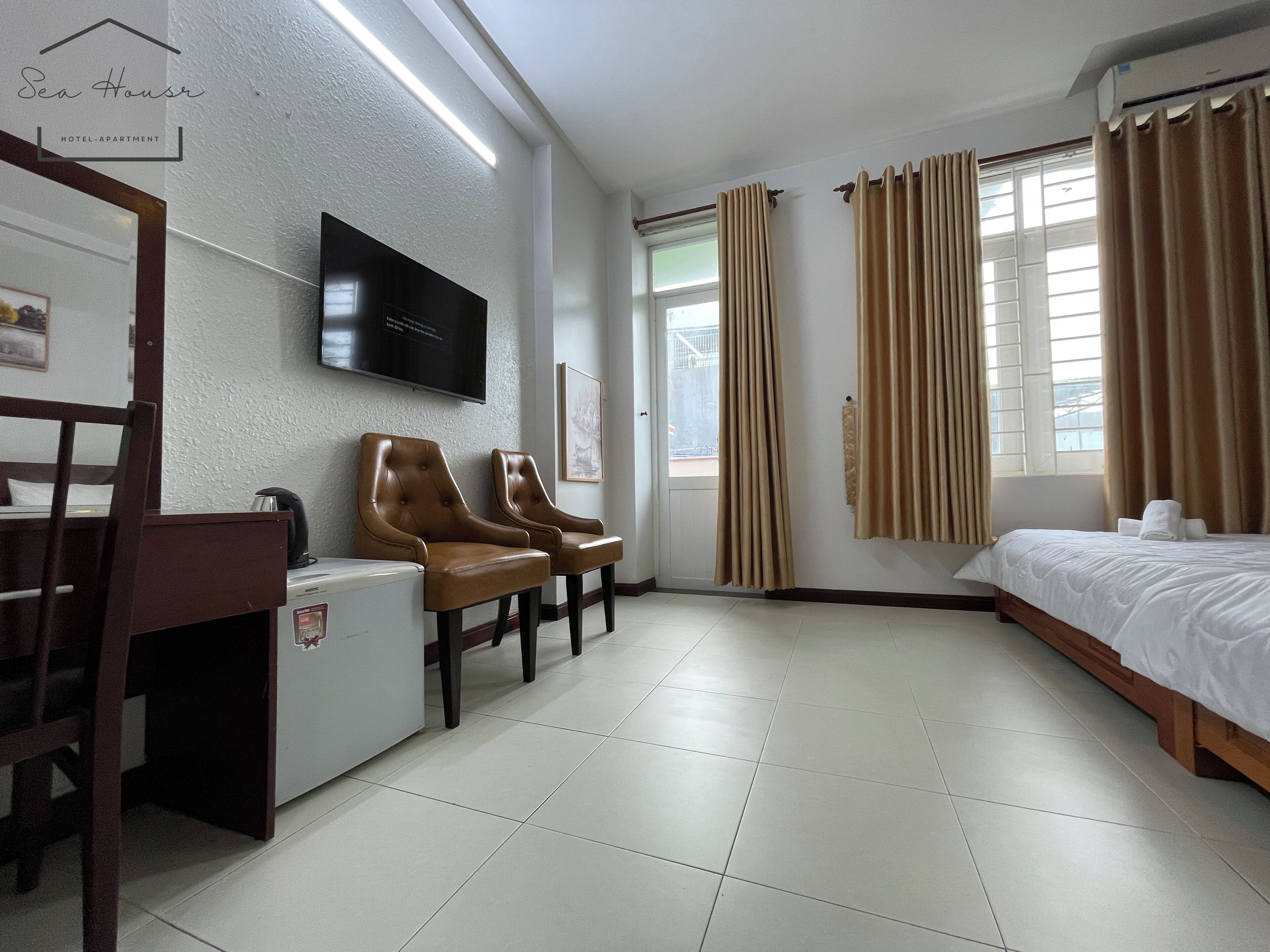Une chambre d’hôtel avec un lit, deux fauteuils, un petit bureau avec une bouilloire, un téléviseur fixé au mur et une fenêtre avec des rideaux.