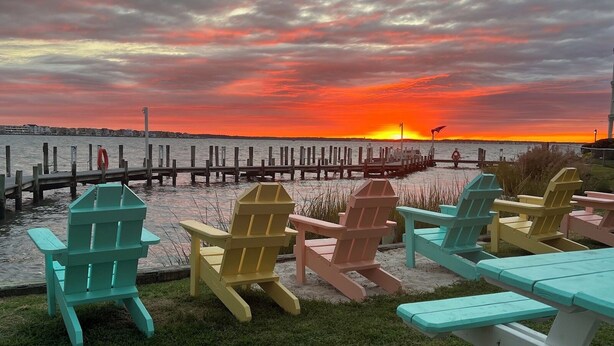 Vista al agua con atardecer, muelle de madera y sillas Adirondack de colores.
