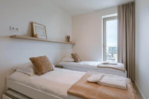 Une chambre d’hôtel avec deux lits, une fenêtre avec des rideaux, une étagère avec un tableau et une lampe, et une vue sur la ville.