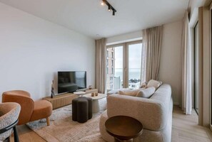 A modern living room with a sofa, ottomans, a TV, and a view of the city through large windows. - Balcony Bliss: Sea View Oasis w/ Free Parking (Oostende)
