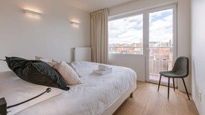 A hotel room with a bed, a chair, a window with a view of buildings, and a ceiling light. - Trendy apartment close to the coast (Oostende)